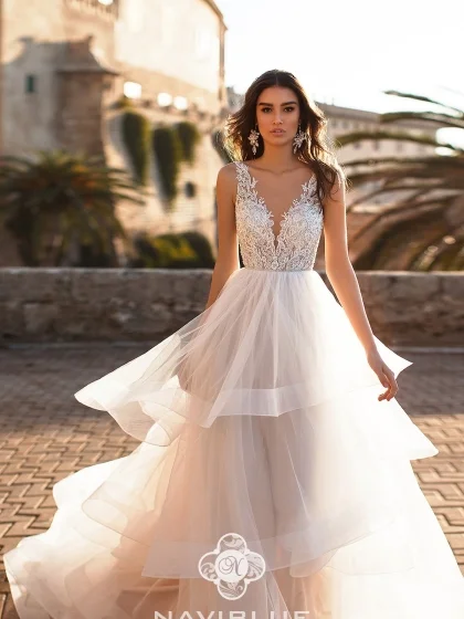 JANELLE 17327 1 Naviblue Современное свадебное платье сочетает одном платье и модную иллюзию прозрачности и игривую пышность в виде воланов, которая в каждом сезоне привлекает смелых современных невест. Юбка в платье многослойная и объемная полностью покрыта оборками по всей длине, верх платья с V-образным вырезом, оформленным цветочным кружевом, делает акцент на тонкую шею и грациозные плечи невесты.