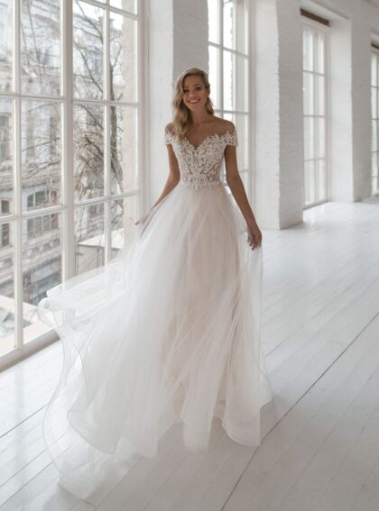 Kamilla - Наталья Романова Kamilla 1 Наталья Романова Потрясающее свадебное платье с блестящей пышной юбкой создает незабываемый, сногсшибательный образ невесты. Многослойная юбка из тюльмарина с пышным шлейфом сверху декорирована кружевным узором. Слегка приспущенные с плеч кружевные элементы романтично закрывают верх платья с фигурным округлым декольте.