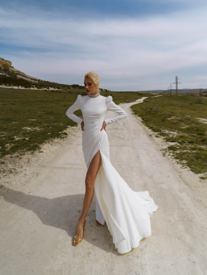 Лея 1 Наталья Романова Свадебное платье прямого силуэта с длинными рукавами и спинкой на пуговичках. Декорировано кристаллами. На юбке разрез по линии бедра.
