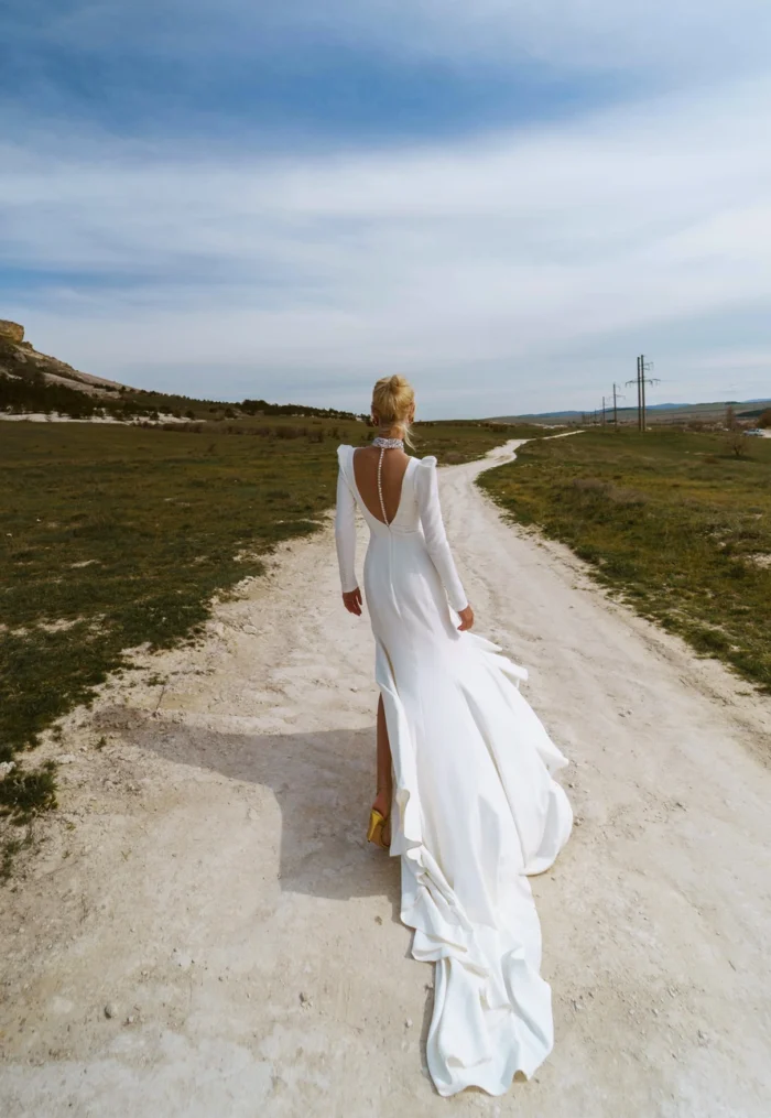 Лея 2 Наталья Романова Свадебное платье прямого силуэта с длинными рукавами и спинкой на пуговичках. Декорировано кристаллами. На юбке разрез по линии бедра.
