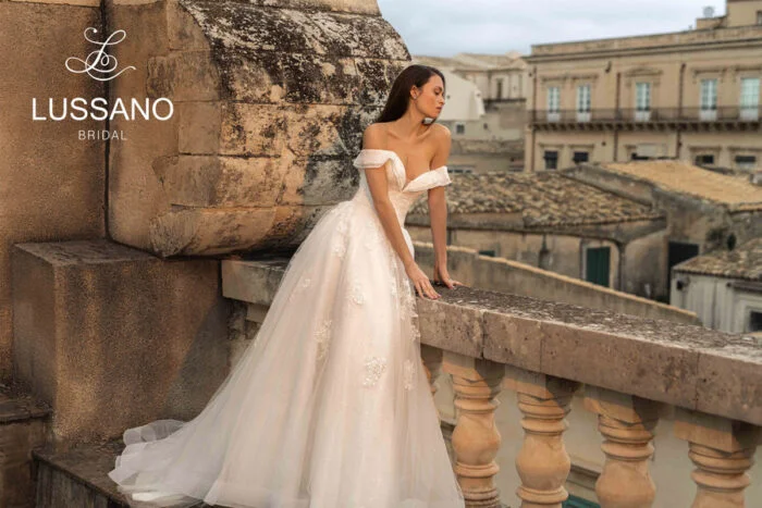 Rosemary 21042 2 Lussano Свадебное платье силуэта принцесса с мерцающей юбкой и утонченным декором. Корсет со шнуровкой на спине и вырезом «сердечко» дополняют спущенные бретели, открывающие плечи. Верх платья декорирован кружевными аппликациями, а талия акцентирована тонким поясом. Многослойная пышная юбка выполнена из фатина, покрытого мелкими переливающимися блестками.