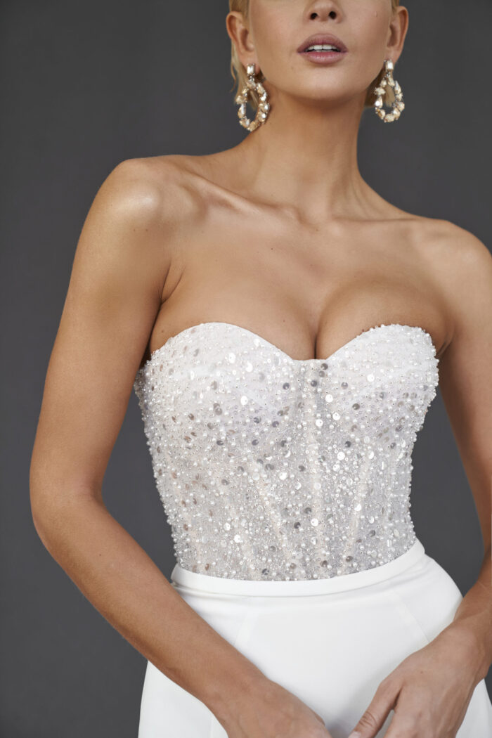 Руби - Martha (Марта) Руби 5 Martha (Марта) Свадебное платье облегающего силуэта — идеальный вариант для невесты, стремящейся подчеркнуть свою фигуру и создать запоминающийся образ. Выполненное из качественного крепа, платье дарит комфорт и свободу движений на протяжении всего дня. Лиф платья изысканно расшит пайетками, которые ловят свет и создают эффектный блеск, добавляя наряду нотку роскоши и элегантности. Тщательно проработанные детали делают этот лиф настоящим произведением искусства, подчеркивающим красоту и женственность. Спинка на молнии обеспечивает отличную посадку и удобство, позволяя невесте уверенно двигаться. Платье плавно обхватывает фигуру, акцентируя внимание на изгибах, а используется мягкий и струящийся креп создает легкость и гармонию в образе.