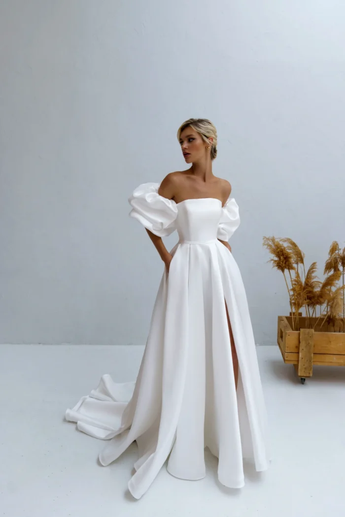 Калипсо 1 Наталья Романова <p style="text-align: justify;">Свадебное платье А-силуэта из атласа с разрезом на юбке и спинкой на молнии идеально сочетает элегантность и современность. Объемные рукава добавляют романтический шарм, подчеркивая изящность силуэта.</p>
